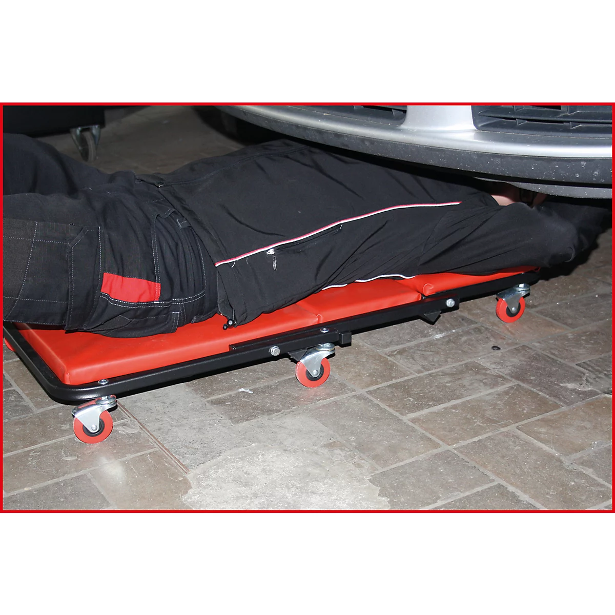 Eine Person liegt auf einer roten Werkstattliege unter einem Auto, um es zu reparieren.